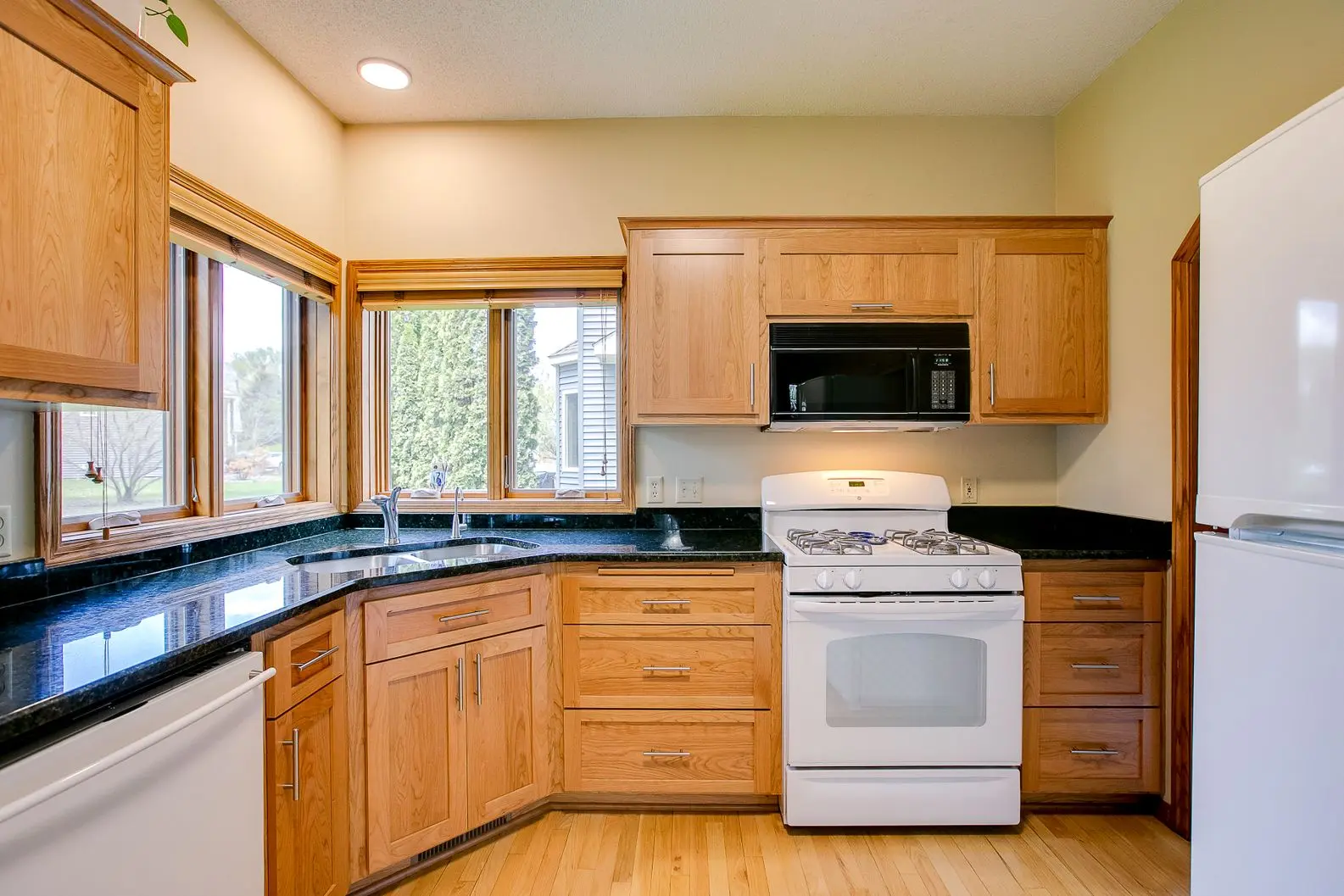 Natural oak cabinets with dark granite countertops and white appliances in a bright kitchen.