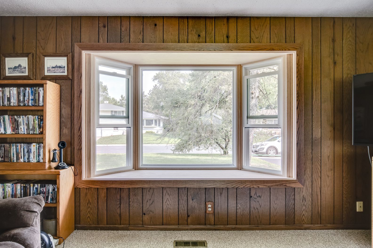Large white bay window with natural wood trim installed in a living room with wood paneling.